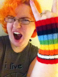 A woman with bright orange hair, wearing glasses, poses with her fist with little and index fingers extended in the "metal" gesture, wearing rainbow knit handwarmers.