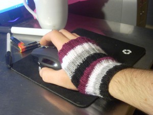 A hand resting on a computer mouse. The hand is wearing a handwarmer knit in the asexual flag colours of black, grey, white and purple.